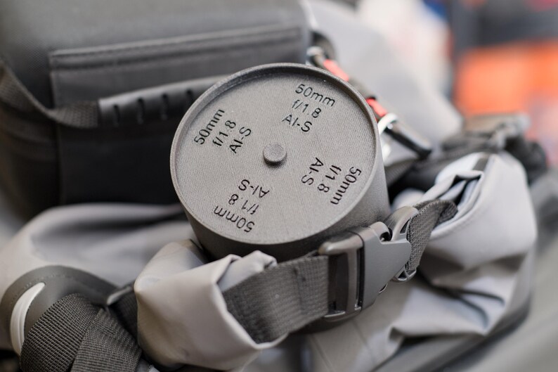 Pode incluir: Uma tampa de lente cinza com o texto "50mm f/1.8 AI-S" est&aacute; presa a uma bolsa de c&acirc;mera. A tampa da lente &eacute; cil&iacute;ndrica com um pequeno bot&atilde;o na parte superior. A bolsa tem uma al&ccedil;a cinza e uma fivela preta.