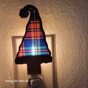 Puede incluir: Una luz de noche decorativa con forma de sombrero de gnomo. El sombrero presenta un patrón de cuadros en azul, naranja y verde, delineado en negro. La luz de noche está enchufada a un enchufe de pared blanco. El texto "Galaxy S21 Ultra 5G" es visible.