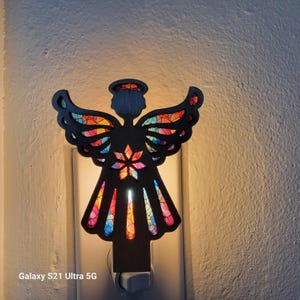 May include: A black angel-shaped night light with stained glass-style wings and dress. The wings and dress feature colorful, geometric patterns. The night light is plugged into a wall outlet and emits a soft glow.