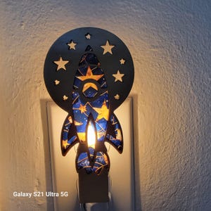 May include: A decorative night light shaped like a rocket ship, with a starry sky design in blue and gold. The rocket is set against a black circular backdrop with star cutouts. The night light is plugged into a wall outlet.