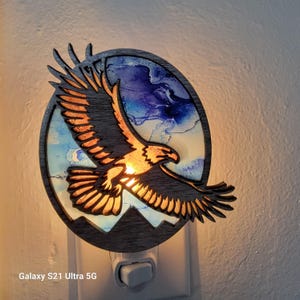 Puede incluir: Una luz de noche de madera con un diseño de águila volando. El águila se representa con las alas extendidas sobre un fondo de arte abstracto azul y blanco. La luz de noche está diseñada para enchufarse a una toma de corriente estándar.