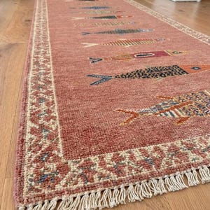 May include: A long, rectangular runner rug in a dusty rose color featuring a repeating pattern of stylized fish in various colors and textures. The rug has a cream-colored border with a geometric design and white fringe at the ends. Text reads ARRUGESTUDIO.
