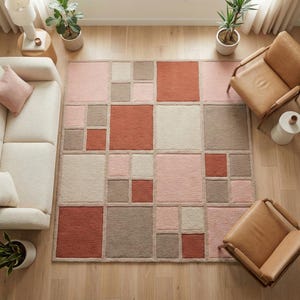 May include: A geometric area rug with a modern design. The rug features squares in shades of pink, beige, and rust, creating a color-blocked pattern. The rug is placed on a hardwood floor, and is surrounded by furniture and plants.