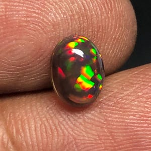 Puede incluir: Una piedra preciosa de ópalo negro de forma ovalada con una superficie lisa y pulida, que presenta destellos vibrantes de rojo, naranja y verde en todo el cuerpo oscuro y translúcido de la piedra, con un corte cabujón abombado.