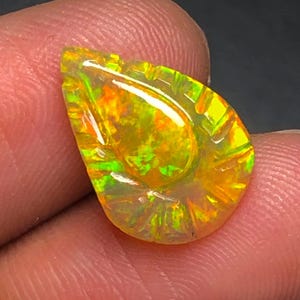 May include: A pear-shaped, carved gemstone featuring a vibrant play of color with dominant orange, yellow, and bright green flashes. The surface of the stone displays intricate, radial grooves and a polished finish that reflects light across its surface.