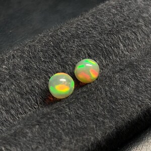 Può includere: Due pietre preziose di opale rotonde con gioco di colori, che mostrano lampi di verde, arancione e giallo. Gli opali sono posti su uno sfondo scuro e strutturato, evidenziando le loro qualità iridescenti. Ideali per la creazione di gioielli.