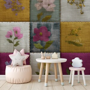 May include: A patchwork wall featuring floral patterns in pink, yellow, and teal. A small white table with wooden legs and a matching chair stand before the wall. A pink ottoman and star-shaped cushions complete the scene.