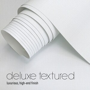 May include: A roll of white, textured wallpaper with a luxurious, high-end finish. The words "deluxe textured" and "luxurious, high-end finish" are printed in black on the wallpaper.