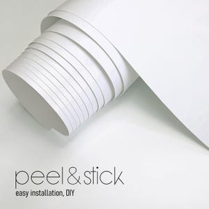 May include: A roll of white peel and stick wallpaper. The roll is partially unrolled, showing the layers of paper. The text "peel & stick" and "easy installation, DIY" are printed in black at the bottom of the image.