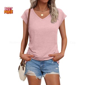 May include: A light pink V-neck t-shirt with short sleeves. The shirt is paired with denim shorts. A gold necklace and a shoulder bag with a brown strap are also visible. The image includes a "BEST PRICE" logo.