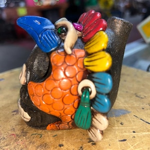 Puede incluir: Una escultura de cerámica de color marrón oscuro con forma de pájaro, con pico azul, pecho texturizado naranja y una cola colorida en forma de abanico con plumas rojas, amarillas, azules, verdes y crema. El pájaro tiene un pequeño ojo verde.