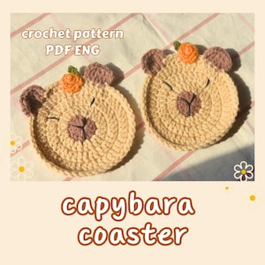 Op de afbeelding: Twee handgemaakte gehaakte capybarat coasters. Elke coaster is rond, beige en heeft een capybaragezicht met bruine oren, een neus en een kleine oranje bloem. De tekst "capybara coaster" wordt weergegeven in bruine letters.