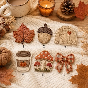 May include: Six embroidered patches featuring autumn themes: a rust-colored maple leaf, a brown acorn, a beige cable-knit sweater with a heart, a coffee cup with a pumpkin icon, a cluster of red-capped mushrooms, and a plaid bow with a brown center.