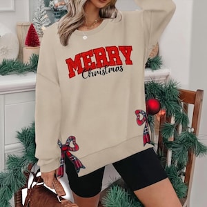 May include: Beige Christmas sweatshirt with the text "MERRY Christmas" in red. The sweatshirt has red plaid bows on the sides. The model is wearing black shorts.