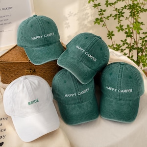 May include: Four baseball caps are displayed. Three caps are dark green with a distressed finish and feature the white embroidered text HAPPY CAMPER. One cap is solid white with the green embroidered text BRIDE on the front panel.