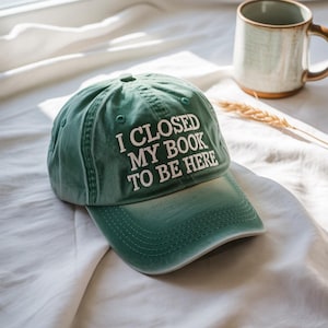 Może przedstawiać: Zielona czapka bejsbolowa z białym haftem "I CLOSED MY BOOK TO BE HERE". Czapka wykonana jest z miękkiej tkaniny i ma zakrzywiony daszek. W tle znajduje się ceramiczny kubek.