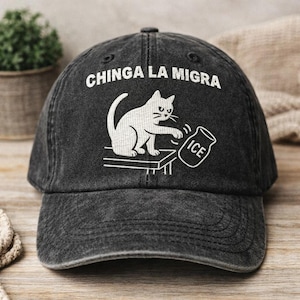 Puede incluir: Gorra de béisbol negra desgastada con el texto "CHINGA LA MIGRA" en blanco. Se presenta una ilustración de un gato blanco, derribando un tarro etiquetado "ICE". La gorra está hecha de un material suave y desgastado.
