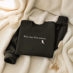 May include: Dark green sweatshirt with the text "Well the f*ck aware." in white, along with a small white graphic. The sweatshirt is displayed on a white, textured surface, with a lit candle and decorative gold accents.