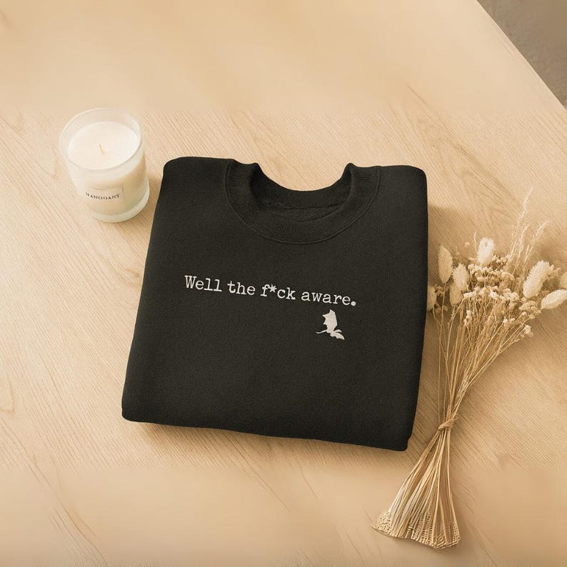 May include: Black crew neck sweatshirt with the text "Well the f*ck aware." in white. A small white graphic of a bird is below the text. A lit candle in a glass jar and a bundle of dried flowers are also in the image.