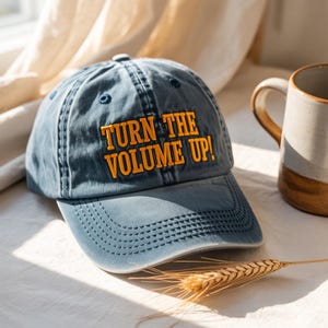 Könnte beinhalten: Eine blaue Baseballkappe mit dem orangefarbenen Text "TURN THE VOLUME UP!". Die Kappe hat einen gebogenen Schirm und einen Knopf oben. Eine Keramiktasse und Weizenähren sind im Hintergrund zu sehen.
