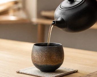 Könnte beinhalten: Eine mattschwarze Keramik-Teekanne gießt Tee in eine abgerundete Keramiktasse mit einer texturierten Verlaufsoptik, die von dunklem Anthrazit an Boden und Rand zu einer gesprenkelten, metallisch bronzefarbenen Mitte des Gefäßes übergeht.