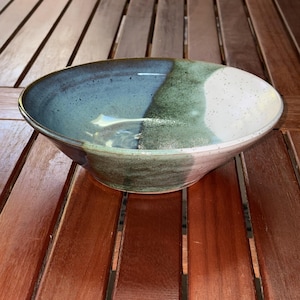 May include: A handmade ceramic bowl with a unique color scheme. The bowl features a gradient of colors, including blue, green, and white. The bowl is round with a wide opening and a slightly textured surface. The bowl is sitting on a wooden table.