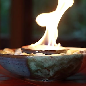 May include: A ceramic bowl with a flame burning in the center. The bowl is filled with small, smooth stones. The flame is bright and yellow, and the bowl has a blue and brown glaze. The bowl is sitting on a red surface.