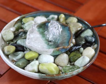 May include: A small, decorative water fountain with a ceramic bowl. The bowl is filled with smooth, colorful stones in shades of green, white, and black. Water flows from a central, light-colored stone feature. The fountain is set on a wooden surface.