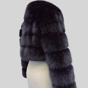 May include: A black fur bolero jacket. The cropped jacket is displayed on a white mannequin. The fur is layered horizontally, creating a textured appearance. The sleeves are long and the jacket is a solid black colour.