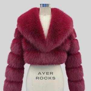 May include: A burgundy faux fur cropped jacket with a wide collar and horizontal banded sleeves. The jacket is displayed on a dress form with the text "AYER ROCKS" printed on the front.