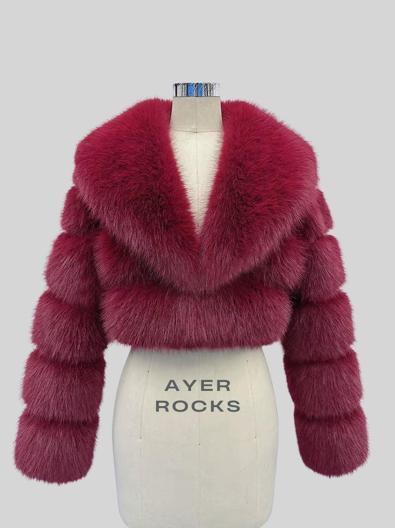 May include: A burgundy faux fur cropped jacket with a wide collar and horizontal banded sleeves. The jacket is displayed on a dress form with the text "AYER ROCKS" printed on the front.