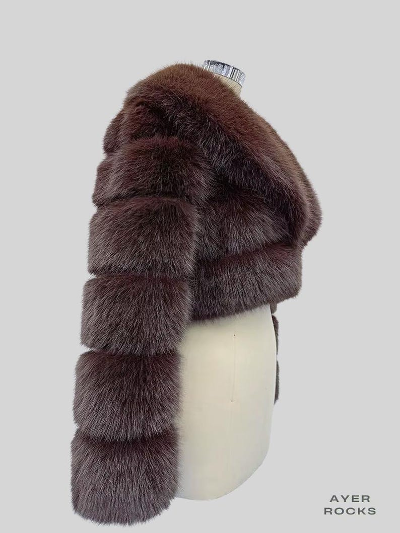 May include: A brown fur jacket with a cropped design. The jacket features horizontal bands of fur, creating a textured appearance. The sleeves are long and the jacket is displayed on a mannequin.