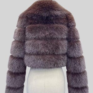 May include: A cropped, brown fur jacket with a horizontal banded design. The jacket has a round collar and long sleeves. The fur appears soft and plush, with a slightly iridescent sheen. The jacket is displayed on a white mannequin.