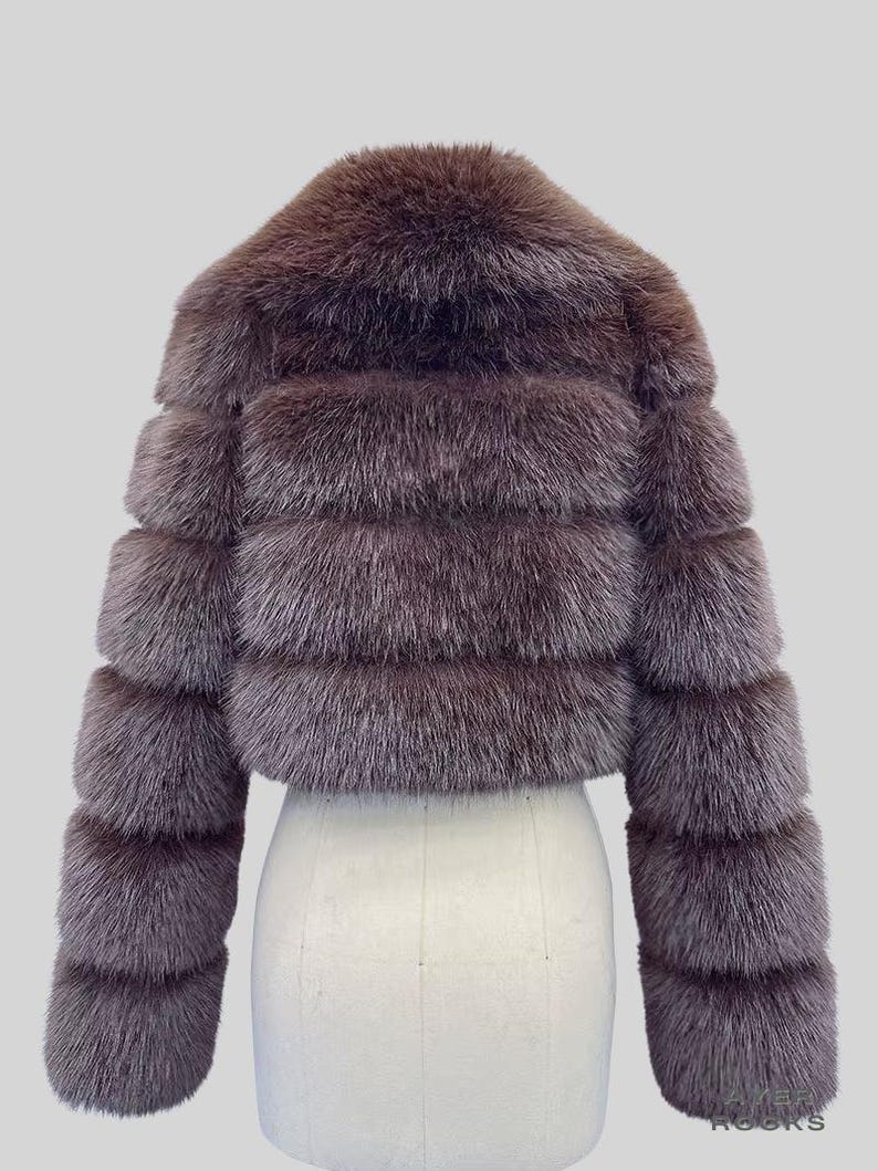 May include: A cropped, brown fur jacket with a horizontal banded design. The jacket has a round collar and long sleeves. The fur appears soft and plush, with a slightly iridescent sheen. The jacket is displayed on a white mannequin.