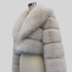 May include: An ivory-coloured faux fur jacket, cropped in style. The jacket has a wide collar and horizontal panels, giving it a textured look. It's displayed on a mannequin against a plain background. The brand name "AYER ROCKS" is visible in the bottom right corner.