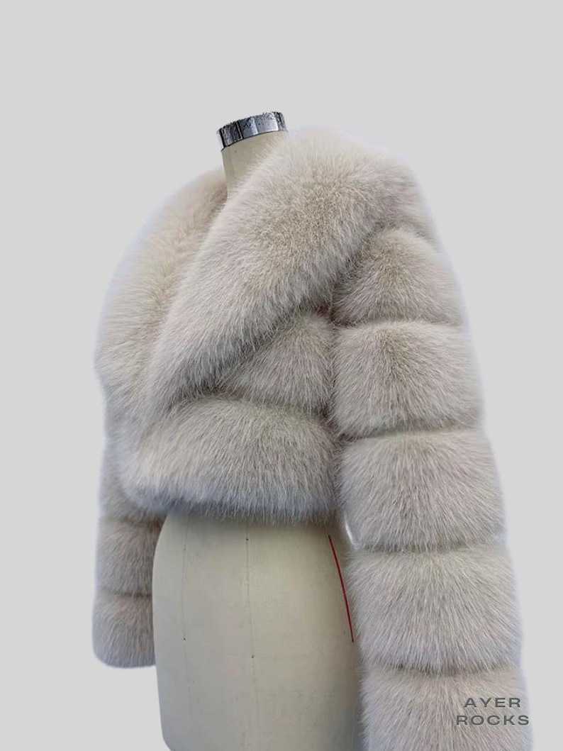 May include: An ivory-coloured faux fur jacket, cropped in style. The jacket has a wide collar and horizontal panels, giving it a textured look. It's displayed on a mannequin against a plain background. The brand name "AYER ROCKS" is visible in the bottom right corner.