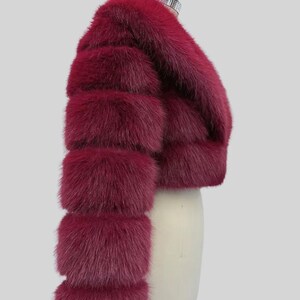 May include: A deep burgundy fur bolero jacket with a cropped design. The jacket has horizontal fur bands on the sleeves and a rounded neckline. The fur looks soft and plush, with a slight sheen. The jacket is displayed on a mannequin.