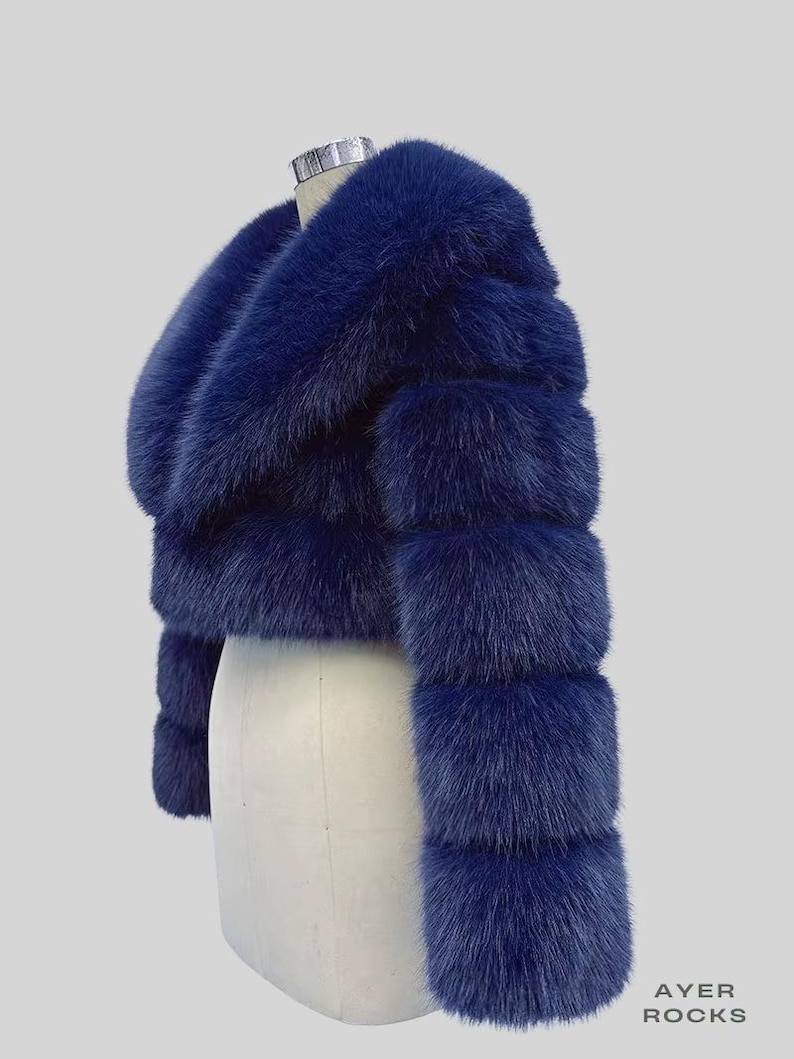 May include: A navy blue faux fur cropped jacket. The jacket has a wide collar and sleeves with horizontal bands. It is displayed on a mannequin against a light grey background. The text "AYER ROCKS" is visible in the lower right corner.