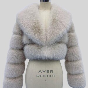 May include: A light grey faux fur jacket, cropped in length, with a broad collar and horizontally banded sleeves. The jacket is displayed on a dress form, with the text "AYER ROCKS" visible. The design and texture suggest a focus on outerwear fashion.