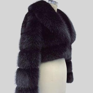 May include: A black fur bolero jacket with a wide collar and three-quarter sleeves. The jacket is displayed on a mannequin against a light grey background. The fur appears soft and luxurious.