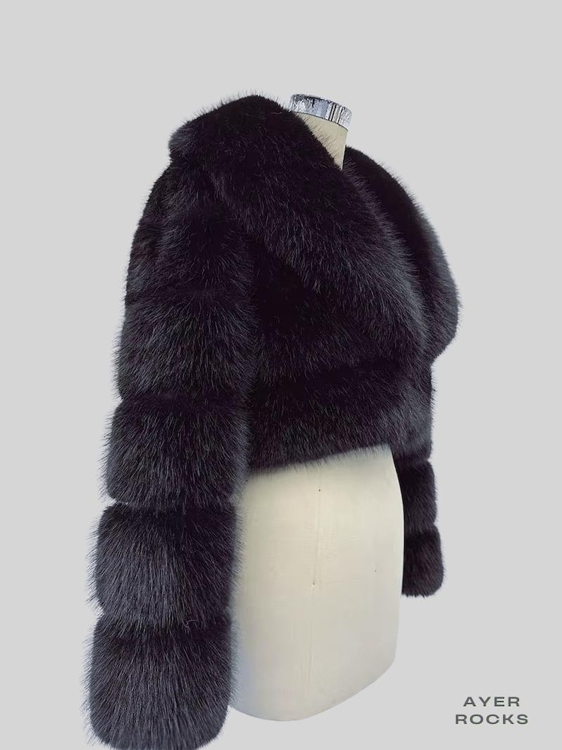 May include: A black fur bolero jacket with a wide collar and three-quarter sleeves. The jacket is displayed on a mannequin against a light grey background. The fur appears soft and luxurious.