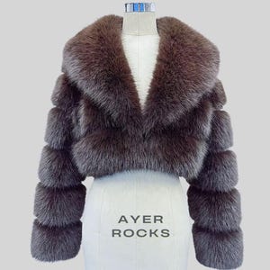 May include: A brown fur cropped jacket with a wide collar and long sleeves. The jacket is displayed on a white mannequin with the text "AYER ROCKS" printed on the front. The jacket's sleeves have a layered design.