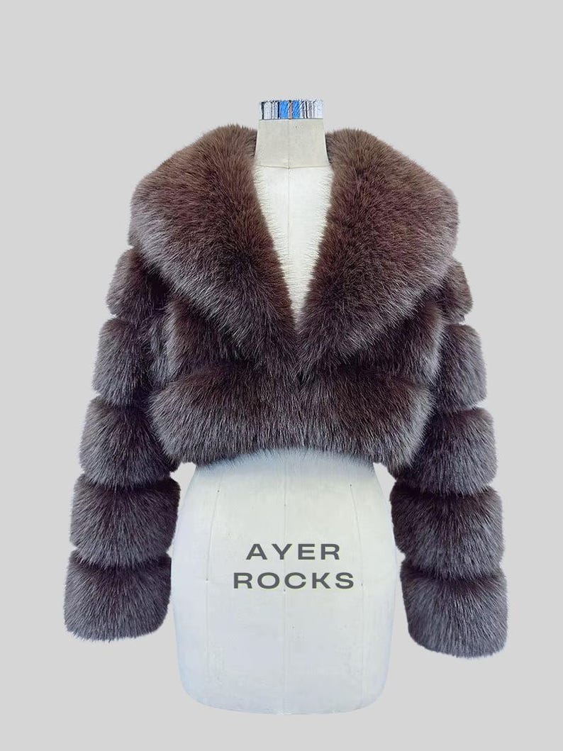 May include: A brown fur cropped jacket with a wide collar and long sleeves. The jacket is displayed on a white mannequin with the text "AYER ROCKS" printed on the front. The jacket's sleeves have a layered design.