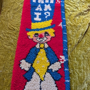 May include: A vintage, handmade rug featuring a colorful clown design. The rug has a red background with a blue border and a blue section at the top with the text "HOW TALL AM I?". The clown has a yellow hat, hair, and diamond-shaped vest.