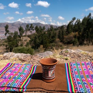 May include: A terracotta-colored drinking vessel with a detailed painted design, set on a brown textile. The textile is adorned with a vibrant, multicolored pattern. The background features a scenic view of mountains and a blue sky.