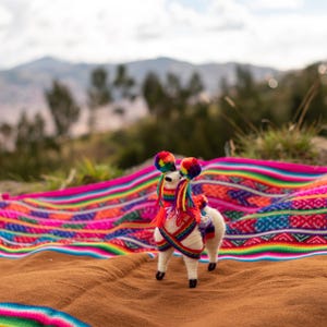 May include: A handcrafted llama figurine with colorful pom-poms and a woven textile. The llama is white with a red and rainbow-colored shawl. The textile has a vibrant pattern of stripes and geometric designs.