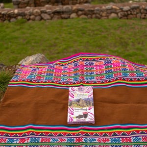 Könnte beinhalten: Ein farbenfrohes gewebtes Textil mit brauner Mitte und lebendigen gemusterten Rändern, mit einer Schachtel Schokolade. Das Textil liegt auf einer Erdoberfläche mit grünem Gras und Steinstrukturen im Hintergrund.