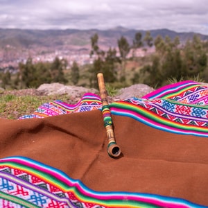 May include: A wooden pipe with a brown bowl and a decorative band of colorful beads rests on a brown blanket with a vibrant, patterned border. The background features a scenic view of a city and mountains.