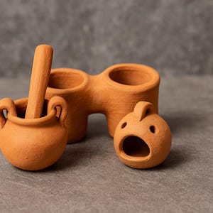 May include: A collection of terracotta pottery pieces. Includes a small mortar and pestle, a double-vessel container, and a small, open-mouthed decorative piece. The items are arranged on a gray surface.