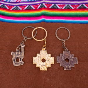Puede incluir: Tres llaveros con colgantes de metal. Uno presenta una llama con el texto "Cusco", otro un diseño de cruz dorada y el tercero un diseño de cruz con figuras. Los llaveros están sobre una superficie marrón con un textil colorido en el fondo.